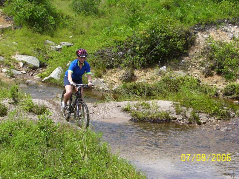 A person in a blue cycling jersey and helmet rides a mountain bike through a shallow stream surrounded by green grass and shrubs. The scene captures a sunny day outdoors, showcasing a natural, rugged landscape with rocks and vegetation. CVI Trails mountain bike trail.