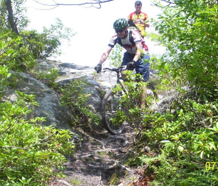 A mountain biker navigates a rocky trail surrounded by lush greenery, while a second person observes in the background. The biker is focused on maneuvering down the steep path, showcasing a challenging biking environment. CVI Trails mountain bike trail.