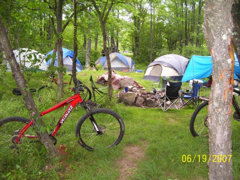 A vibrant camping scene featuring several tents nestled among green trees. In the foreground, two mountain bikes are parked near a fire pit surrounded by rocks, with camping chairs set up nearby. The area is lush with grass and the atmosphere suggests a peaceful outdoor retreat. CVI Trails mountain bike trail.