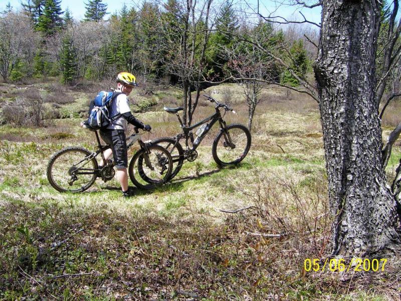 A person in a yellow helmet and backpack stands next to two mountain bikes in a grassy area surrounded by sparse trees and shrubs on a sunny day. The photo captures a peaceful outdoor scene, suggesting an adventure in nature. CVI Trails mountain bike trail.