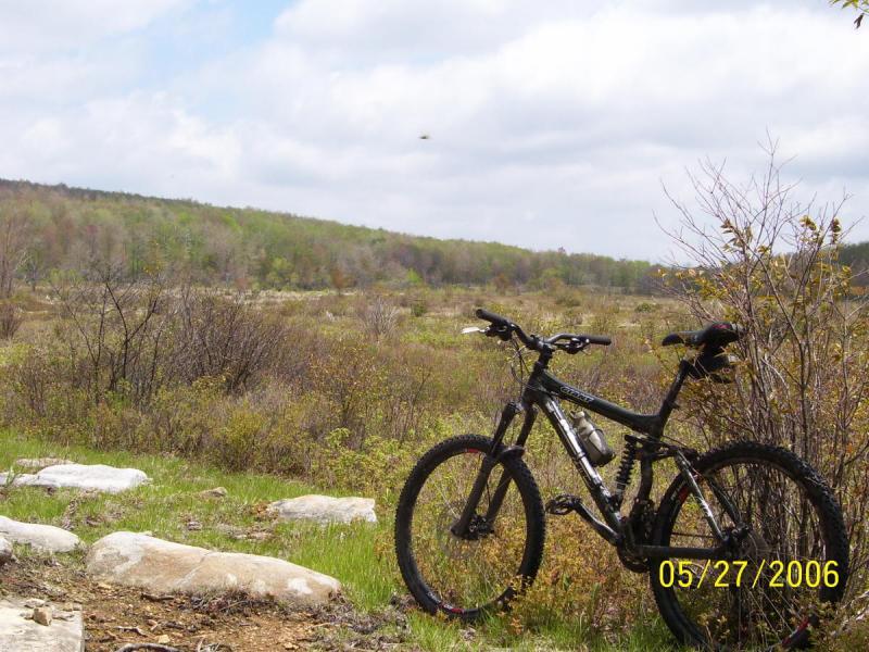 A mountain bike parked on a rocky trail, overlooking a vast landscape of green hills and sparse trees under a cloudy sky. The setting is rural, suggesting an outdoor recreational area, with the bike positioned prominently in the foreground. CVI Trails mountain bike trail.