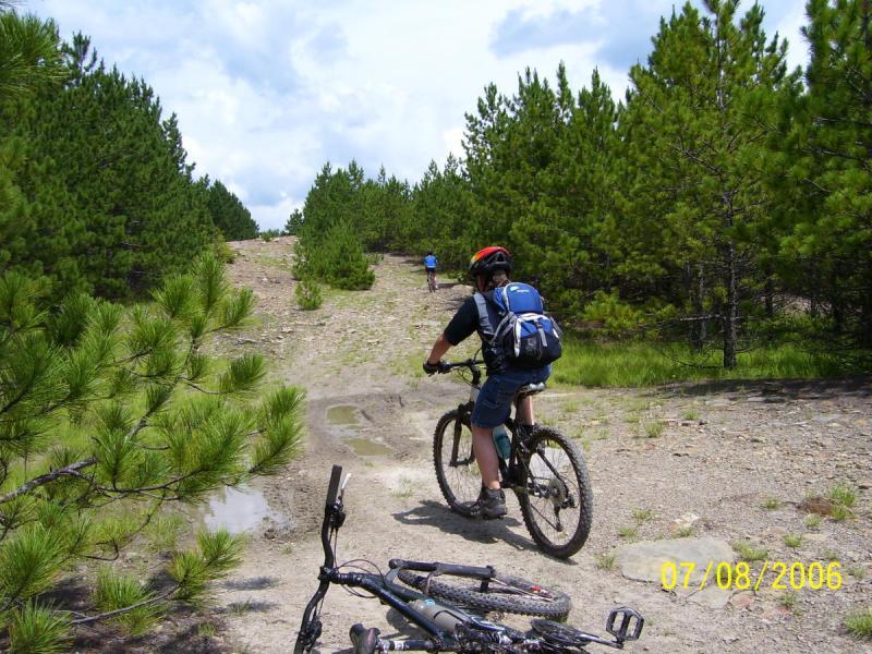 A cyclist riding up a gravel path surrounded by pine trees, with another person walking ahead. In the foreground, a mountain bike rests on the ground, and the scene is set against a partly cloudy sky. CVI Trails mountain bike trail.