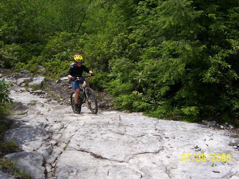 A young person riding a mountain bike on a rocky trail surrounded by lush green vegetation. The rider is wearing a yellow helmet and appears to be navigating a steep section of the path. CVI Trails mountain bike trail.