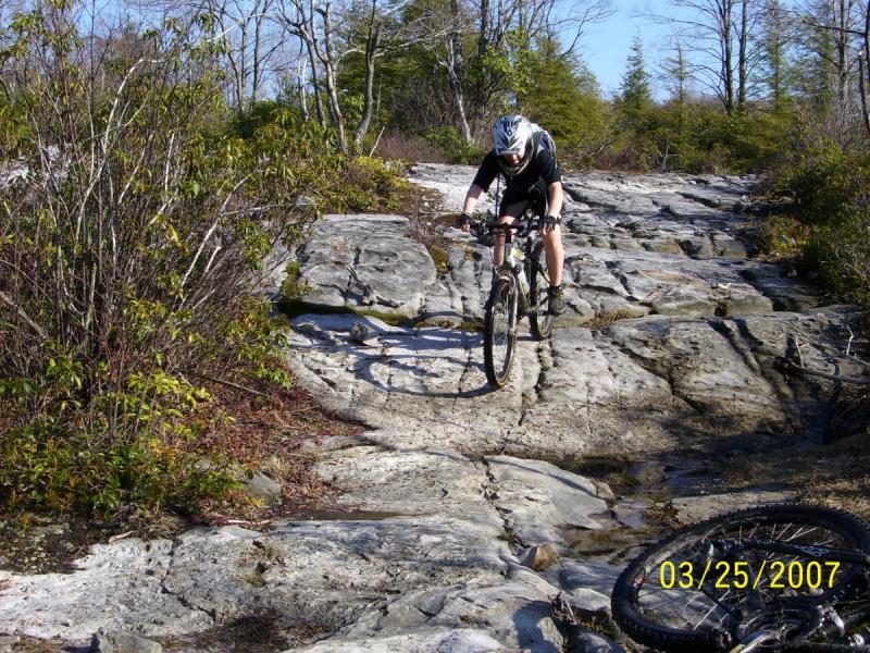 A mountain biker navigating a rocky trail in a wooded area, with a focus on the challenging terrain. The biker is wearing a helmet and cycling gear, demonstrating an active outdoor experience. In the foreground, a second bike is on the ground, suggesting a rugged biking adventure. CVI Trails mountain bike trail.