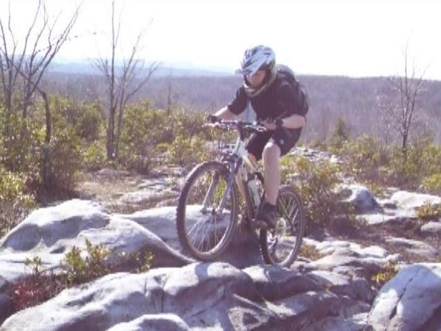 A mountain biker navigating rocky terrain on a sunny day. The rider is wearing a helmet and riding on a bike with both front and back wheels elevated on the rugged rocks. In the background, there are sparse trees and a view of the landscape stretching into the distance. CVI Trails mountain bike trail.