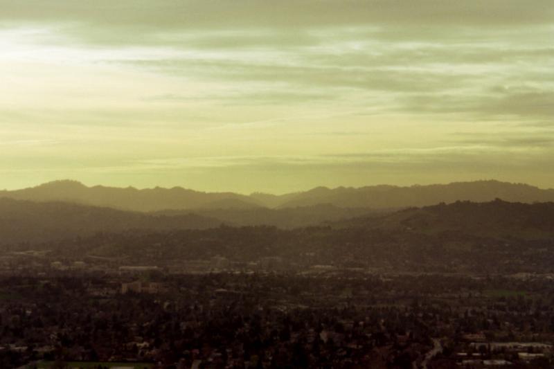 A panoramic view of rolling hills and valleys under a soft, golden sky, with urban structures visible in the foreground. The landscape features a mix of greenery and developed areas, creating a contrast between nature and city life. The overall atmosphere is calm and serene, indicative of either dawn or dusk. Lime Ridge mountain bike trail.