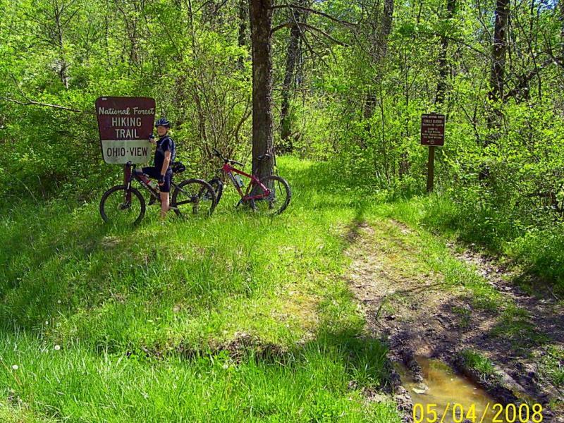 A person stands next to two bicycles by a sign indicating the beginning of the National Forest Hiking Trail, labeled "Ohio-View." Surrounded by lush greenery and tall trees, the trail is visible in the background, leading into the forest. Ohio View Trail mountain bike trail.
