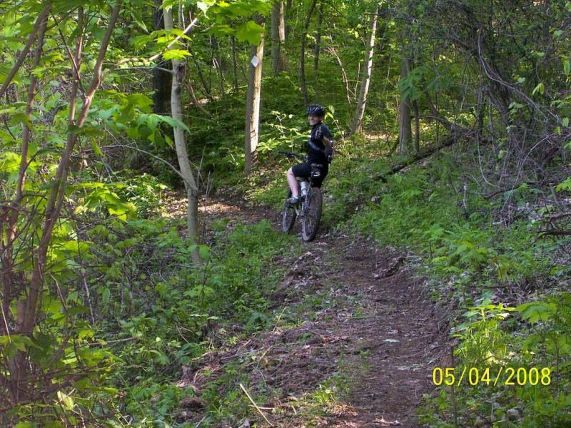 A person in cycling attire sitting on a mountain bike along a dirt trail surrounded by lush green foliage and trees in a forest setting. The scene captures the peacefulness of nature, highlighting the adventure of mountain biking. Ohio View Trail mountain bike trail.