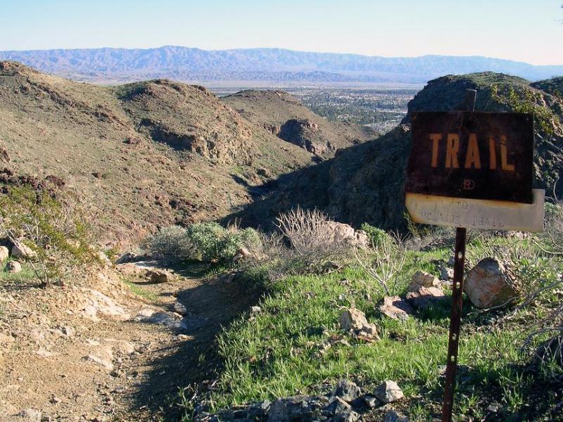 A dirt trail winding through rocky terrain with a weathered sign labeled "Trail" in the foreground, surrounded by sparse vegetation and hills in the background, leading to a panoramic view of distant mountains and a valley. The Goat Trails mountain bike trail.