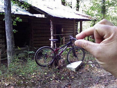 A hand is positioned in the foreground, seemingly pinching a bicycle that is leaning against a wooden cabin in a forested area. The cabin has a metal roof and is surrounded by trees and a dirt ground. Clinton Nature Preserve mountain bike trail.