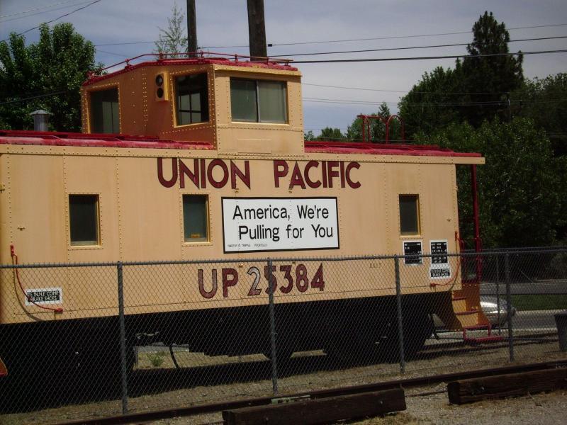 Alt text: A Union Pacific caboose painted in tan and red, featuring a prominent sign that reads "America, We're Pulling for You" along with the number UP 25384. The caboose is surrounded by a fence with trees and power lines in the background. Bizz Johnson Trail mountain bike trail.