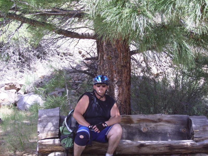 A person sitting on a wooden log in a natural setting, wearing a black sleeveless shirt and blue shorts, a helmet, and sunglasses. The background features a large pine tree and some greenery, indicating a woodland area. The individual appears relaxed, with a cigarette in their mouth. A backpack is placed beside them. Bizz Johnson Trail mountain bike trail.