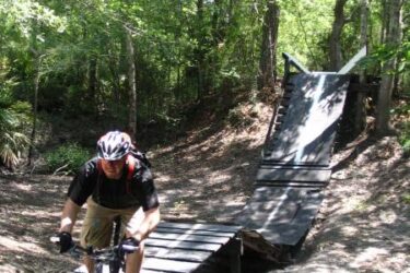 A mountain biker riding on a wooden ramp in a forested area, with trees and greenery surrounding the trail. The ramp leads into a jump, offering an adventurous path for biking enthusiasts. Alapaha Trail mountain bike trail.