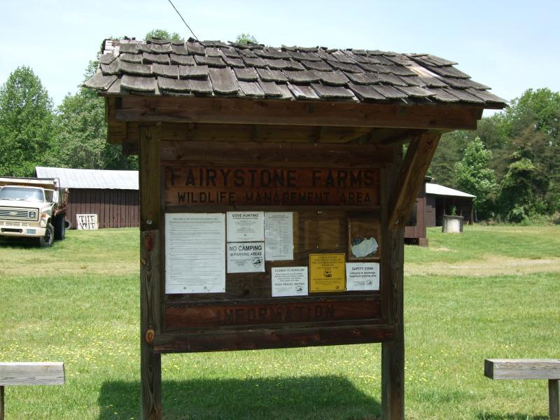 Wooden information board for Fairystone Farms Wildlife Management Area, displaying rules and guidelines. In the background, a vintage truck and rustic buildings are visible, surrounded by grassy fields and trees. Adams Track Trails mountain bike trail.