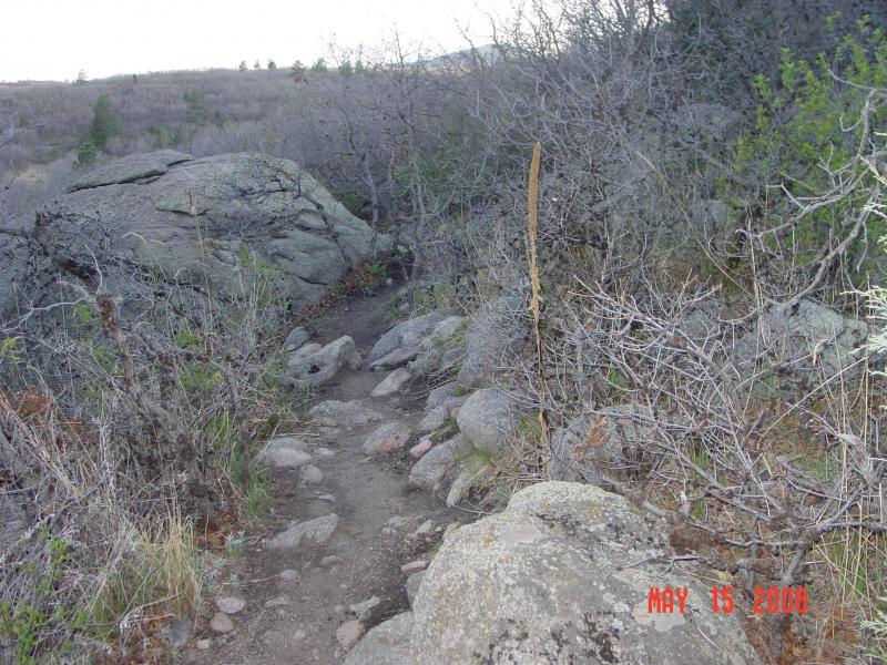 A narrow hiking trail winds through rocky terrain, surrounded by sparse vegetation and scattered boulders. The path appears rugged with loose stones and is lined with dry, leafless shrubs. A wooden marker stands alongside the trail, indicating direction or proximity to a landmark. The background features distant hills and trees, suggesting a natural landscape. Cheyenne Mountain State Park mountain bike trail.