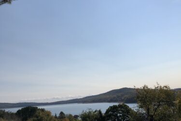 Scenic view of a lake surrounded by greenery, with a viewpoint sign in the foreground providing information about the area. The sky is clear with a bright sun shining. Mountains can be seen in the distance across the water. Round Valley Recreation Area mountain bike trail.
