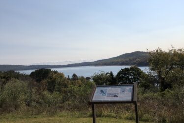 Scenic view of a lake surrounded by hills and trees, with an informational sign in the foreground. The sky is clear, indicating a sunny day. Round Valley Recreation Area mountain bike trail.