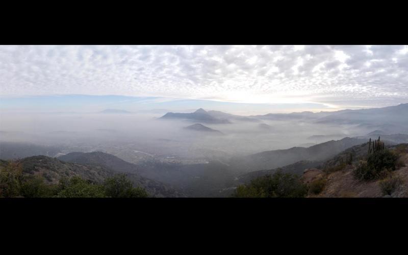 A panoramic view of a mountainous landscape shrouded in mist, with soft clouds scattered across the sky. The scene captures a tranquil atmosphere with layers of hills and valleys visible below, partially obscured by fog. Cancha Carrera mountain bike trail.