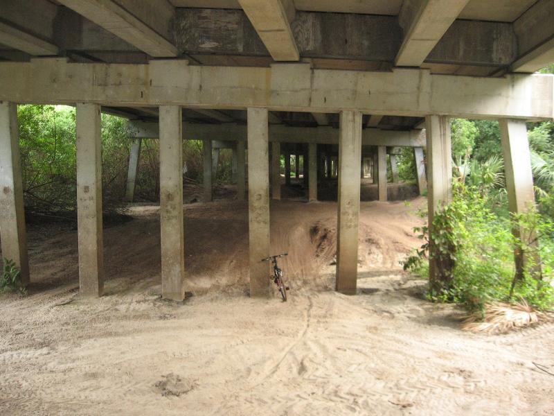 An empty area beneath a concrete bridge, featuring tall support columns and a sandy ground. A bicycle is leaned against one of the columns, surrounded by greenery and dirt mounds. Fort Pierce Mountain Bike Trail mountain bike trail.