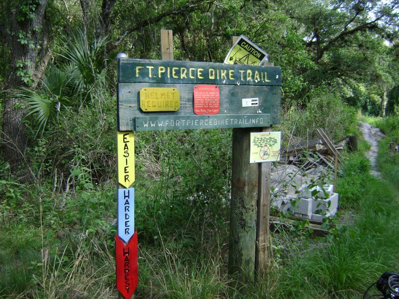Signpost for the Fort Pierce Bike Trail, featuring information on safety regulations, including a helmet requirement. The sign includes difficulty level indicators—marked as easier, harder, and hardest—along with cautionary information and a website link for more details. Surrounding vegetation and a visible bike path are also present in the background. Fort Pierce Mountain Bike Trail mountain bike trail.