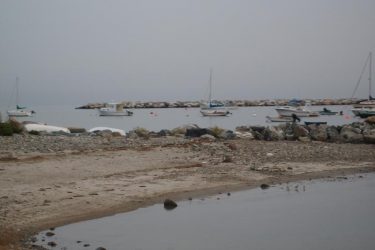 A serene coastal scene featuring several boats anchored in calm waters near a rocky shoreline. In the foreground, a sandy beach meets the water, with scattered pebbles and plants. The background is shrouded in a misty atmosphere, creating a tranquil and slightly overcast ambiance. Hwy 1a From Wallis Sands State Park mountain bike trail.