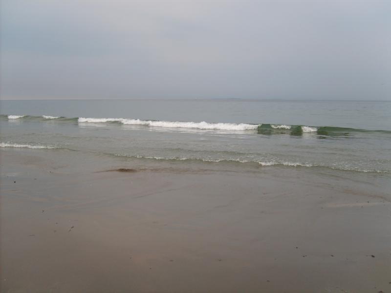 A serene beach scene featuring gentle waves lapping at the shore under a cloudy sky. The sandy beach is visible in the foreground, and the calm ocean stretches into the distance. Hwy 1a From Wallis Sands State Park mountain bike trail.