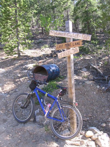 A blue mountain bike leans against a wooden signpost in a forested area, with directional signs pointing towards "Warren-Gulch Road," "Lost Mine Lane," and "Trail." A blue barrel is positioned next to the bike. The surrounding terrain is rugged and natural, with green foliage in the background. Warren Gulch mountain bike trail.