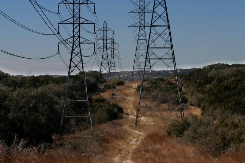 A rural landscape featuring tall power lines and electrical towers alongside a dirt path surrounded by dry grass and sparse vegetation. The scene is set against a clear blue sky, with distant hills visible in the background. Behind The Rim mountain bike trail.