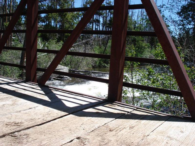 Alt tag: "View from a wooden bridge with a metal railing, overlooking a rushing river surrounded by lush greenery and trees under a clear blue sky." Middle Fork Popo Agie River mountain bike trail.