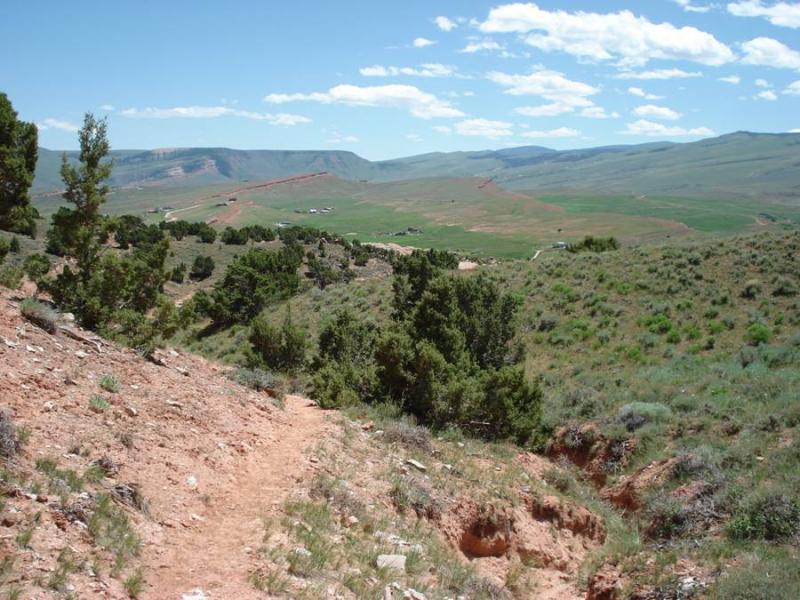 A winding dirt path leads through a green valley surrounded by hills under a clear blue sky with scattered clouds. Sparse trees dot the landscape, and distant structures are visible on the horizon, indicating a rural setting. The Bus mountain bike trail.