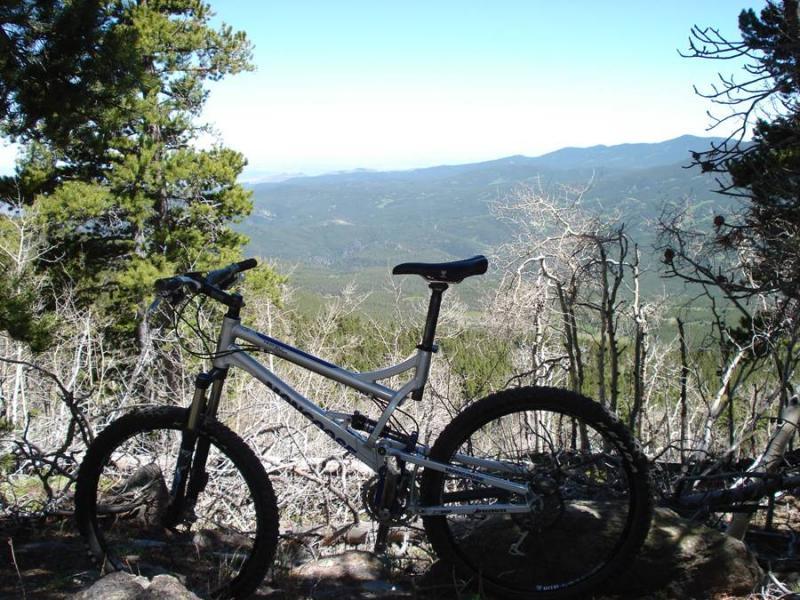 A mountain bike positioned on a rocky ledge with a panoramic view of rolling hills and distant mountains in the background. The scene features lush green trees and a clear blue sky, emphasizing the outdoor adventure setting. White Pine Mountain mountain bike trail.