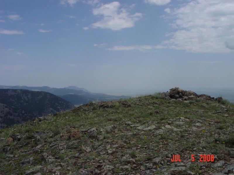 A scenic view from a rocky mountain summit, featuring green grass, scattered rocks, and distant mountain ranges under a partly cloudy sky. The image captures a sense of elevation and natural beauty. The date of the photo is July 5, 2008. Collar Peak mountain bike trail.