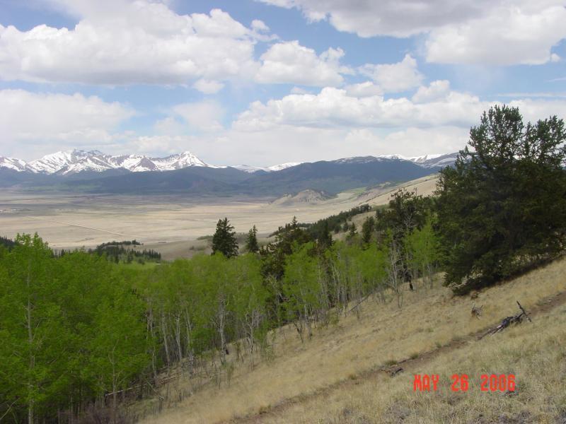 A panoramic view of a mountainous landscape featuring snow-capped peaks under a partly cloudy sky. In the foreground, green trees and grass cover a grassy slope, while a valley stretches out below, leading to distant hills. The scene captures the beauty of nature on a clear day. Colorado Trail: Kenosha Pass To Lost Creek Wilderness Bound mountain bike trail.