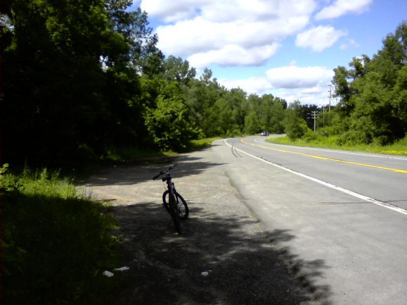 A scenic view of a rural road with a bicycle parked on the shoulder. The background features lush green trees under a partly cloudy sky, indicating a sunny day. The road curves gently in the distance. Lock 18 mountain bike trail.