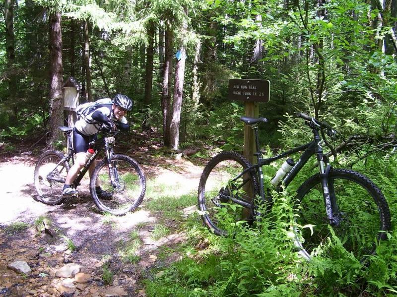 A mountain biker in a cycling outfit leans over their bike at a fork in a wooded trail. Nearby, a second bike is propped against a sign that reads "Red Run Trail" with information about the trail's length. The trail is surrounded by lush green foliage and tall trees, indicating a scenic outdoor environment. Red Run Trail mountain bike trail.