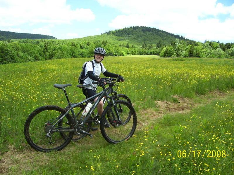 A person wearing a helmet and cycling gear stands next to two mountain bikes in a vibrant green meadow filled with yellow wildflowers. In the background, rolling hills and a clear blue sky are visible, suggesting a sunny day outdoors. Red Run Trail mountain bike trail.