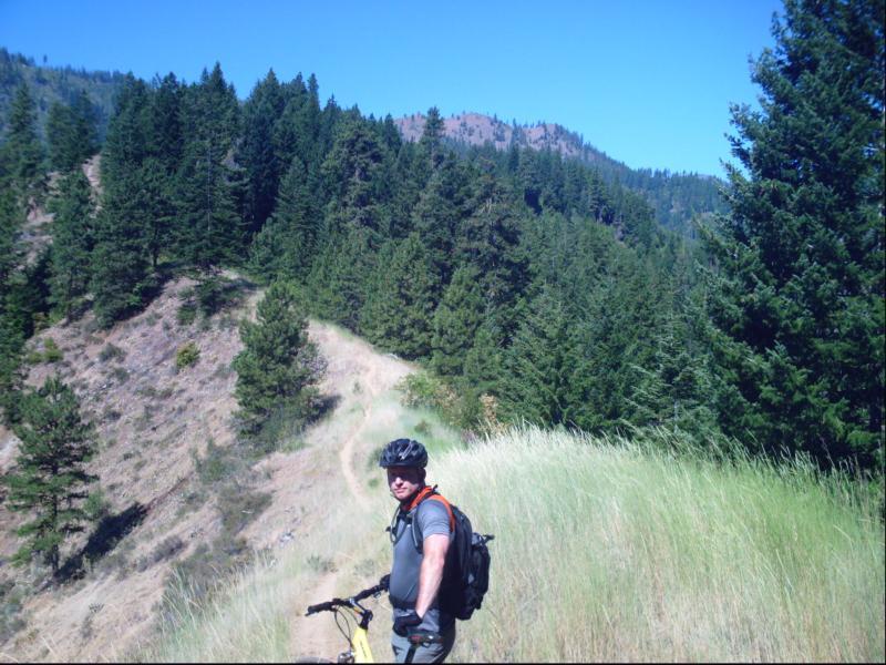 A person in a cycling helmet stands beside a mountain bike on a grassy trail, surrounded by lush green trees and a scenic hill. The sky is clear and blue, indicating a sunny day. Freund Creek mountain bike trail.