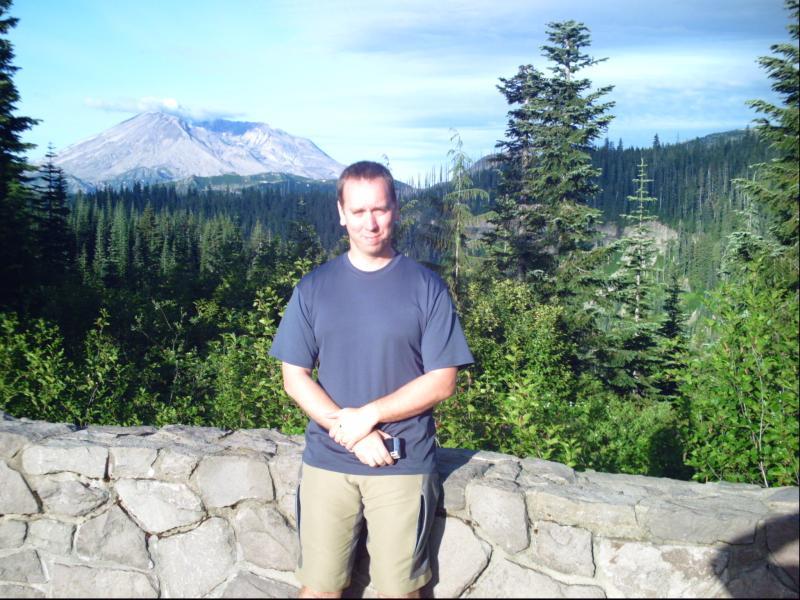 A man stands in front of a stone wall surrounded by lush green trees, with a mountain in the background under a clear blue sky. He is wearing a gray t-shirt and khaki shorts, and appears to be enjoying the scenic landscape. Smith Creek Epic mountain bike trail.