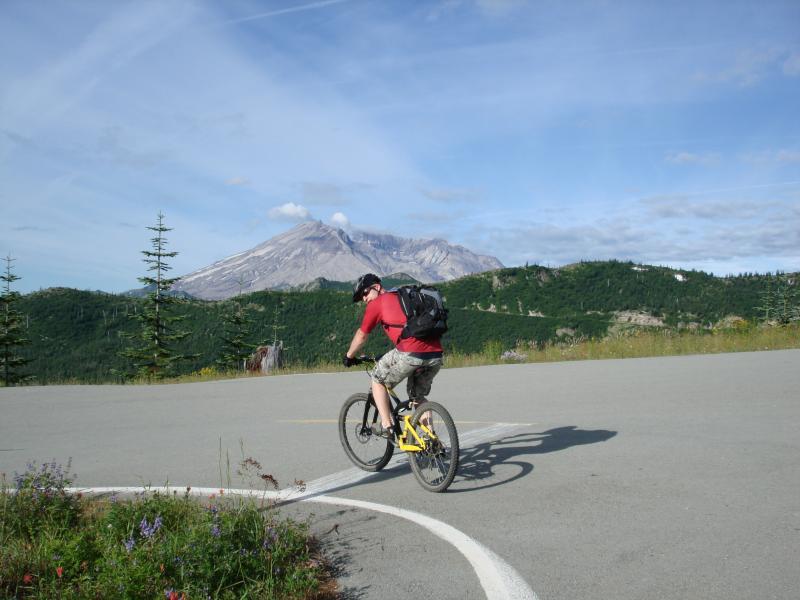 A person riding a mountain bike on a paved path, with a scenic view of a mountain and lush greenery in the background. The cyclist is wearing a red shirt, shorts, and a backpack, and is seen from behind as they navigate a curve in the road. Smith Creek Epic mountain bike trail.