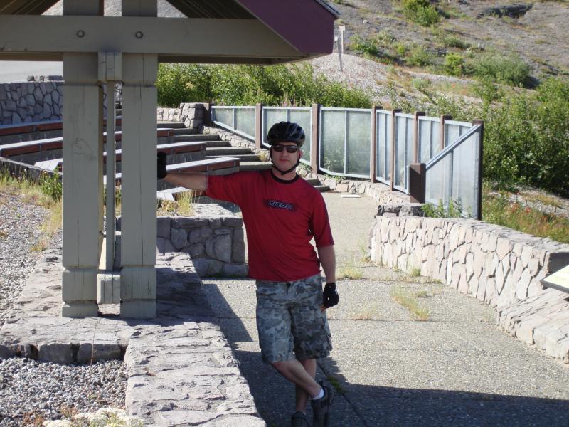 A person wearing a helmet and sunglasses stands casually with one arm resting on a wooden structure, dressed in a red t-shirt and camouflage shorts. The surrounding area features a paved path and grassy hillside, indicating an outdoor location, likely a scenic or recreational site. Smith Creek Epic mountain bike trail.
