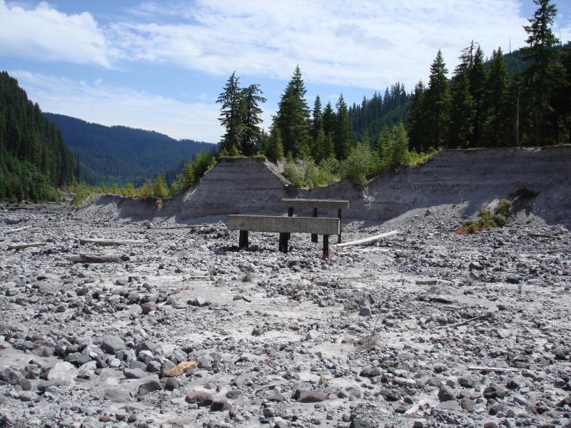 A barren landscape featuring a rocky, gray surface with a wooden platform partially embedded in the ground. Surrounding the platform are tall green trees and steep, eroded banks in the background, all under a blue sky with some clouds. Smith Creek Epic mountain bike trail.
