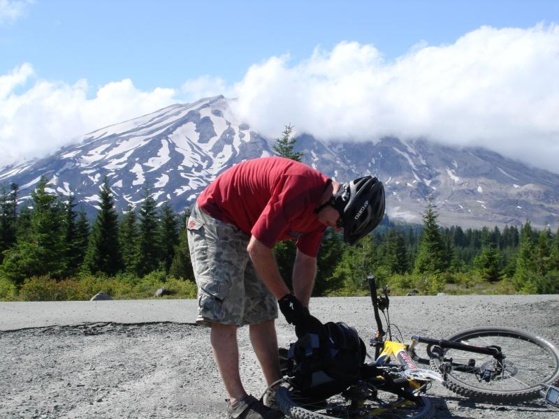 A person wearing a helmet and a red shirt is adjusting equipment near a mountain bike on a gravel path. In the background, a snow-capped mountain rises under a blue sky with scattered clouds, surrounded by lush green trees. Smith Creek Epic mountain bike trail.