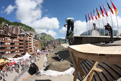 Mountain biker performing a jump off a wooden ramp, with a backdrop of a lively outdoor festival featuring flags from various countries, and a mountain landscape under a blue sky with fluffy clouds.