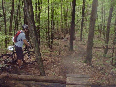 A mountain biker standing next to a bike on a wooded trail surrounded by green trees, preparing to navigate a downhill path. Dufferin County mountain bike trail.