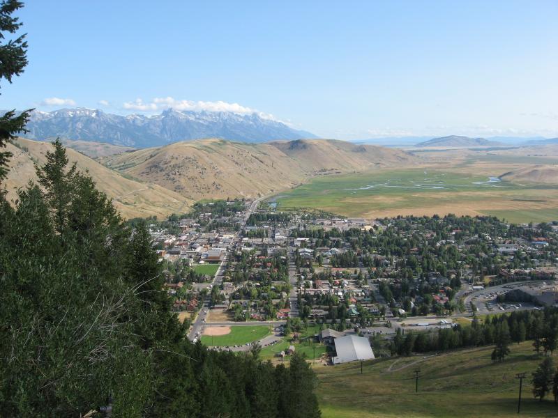 A panoramic view of a small town nestled in a valley, surrounded by rolling hills and distant snow-capped mountains under a clear blue sky. The town features a grid of streets, green trees, and a grassy field, with buildings and homes visible along the landscape. Snow King Mountain mountain bike trail.