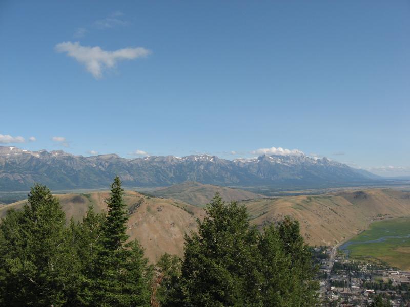 A panoramic view of a mountainous landscape under a clear blue sky, featuring green trees in the foreground, rolling hills, and a distant mountain range with snow-capped peaks. Below, a small town is visible near a winding river. Snow King Mountain mountain bike trail.