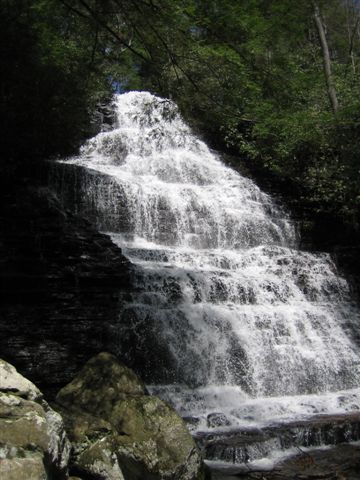 A cascading waterfall surrounded by lush green trees and rocky terrain, with water flowing elegantly over layers of dark stone.