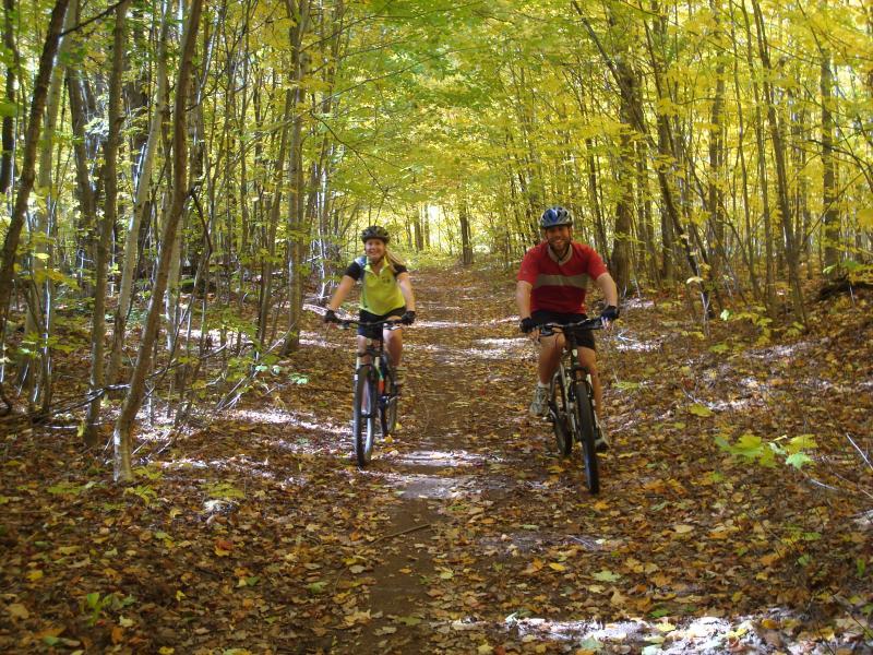 Two people riding bicycles along a dirt path in a forest during autumn. The path is surrounded by trees with vibrant yellow and green leaves, creating a picturesque fall setting. The cyclists are smiling and enjoying the ride, with fallen leaves covering the ground. Noquemanon Trails Network: South Marquette Trails mountain bike trail.