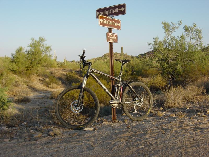A mountain bike parked at a trail junction in a desert landscape, with signs indicating the direction to Moonlight Trail and San Tan Trail. Surrounding vegetation includes cacti and low shrubs, and the ground is a mix of dirt and rocky terrain, under a clear blue sky. San Tan mountain bike trail.