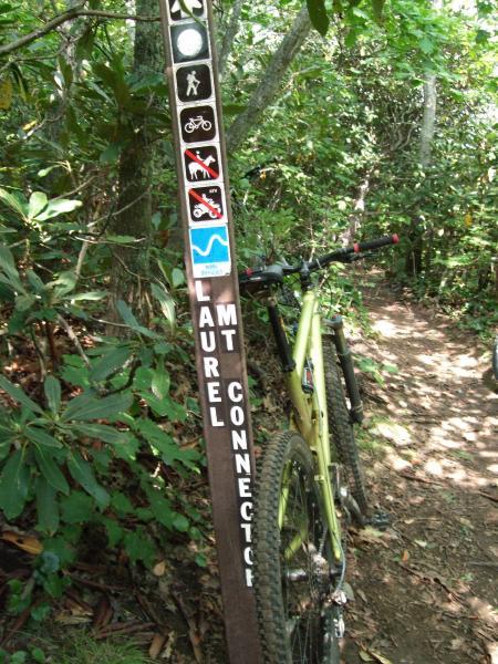 A mountain bike leaning against a trail sign in a dense wooded area. The sign indicates the "Laurel Mountain Connector" and features various symbols, including those for hiking, biking, a no-pets rule, and a trail with a winding path. The trail is narrow and surrounded by lush greenery. Laurel Mountain mountain bike trail.